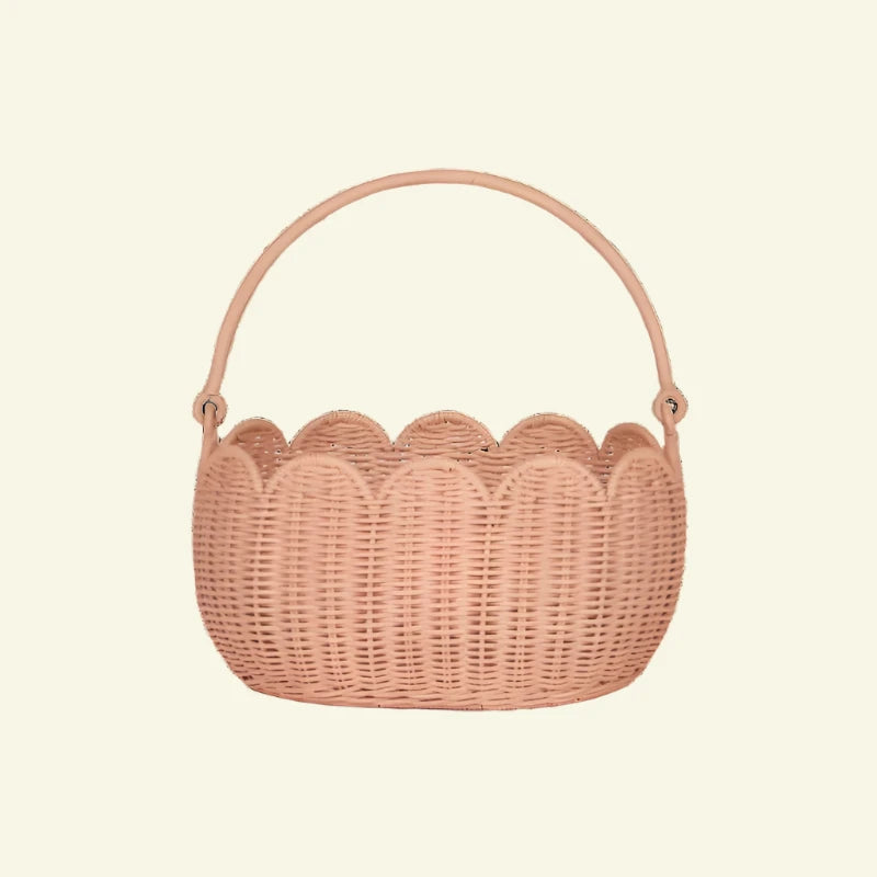 The Olli Ella Petal Basket in Seashell Pink features a scalloped design with a sturdy arched handle, crafted from natural wicker for rustic charm. Perfect for spring adventures or displaying against a neutral backdrop, it seamlessly combines practicality and style.