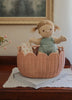 A plush doll with blond pigtails in a mint green outfit sits in an Olli Ella Petal Basket - Seashell Pink, placed on a book over a lace mat on a wooden table. A framed painting and small plant bring charm to this delightful springtime scene.