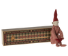 A small doll dressed in a red and white striped knitted suit and a pointed hat sits beside a decorative matchbox labeled "Maileg Long Legged Pixy" from "The Grand Old Match Factory." The doll has a simple, whimsical design with a smiling face, capturing the enchanting pixy style.