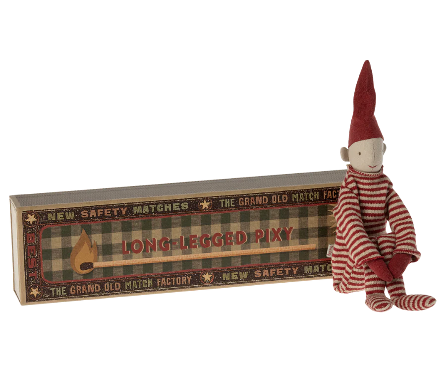 A small doll dressed in a red and white striped knitted suit and a pointed hat sits beside a decorative matchbox labeled "Maileg Long Legged Pixy" from "The Grand Old Match Factory." The doll has a simple, whimsical design with a smiling face, capturing the enchanting pixy style.