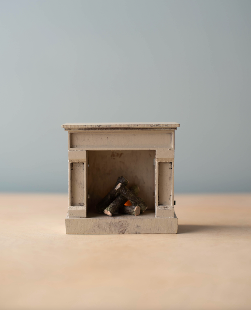 A small, beige miniature fireplace with tiny logs sits on a light surface beside the Maileg Moms Afternoon Tea Set, against a plain, light blue background. Choking hazard—keep out of reach of young children.