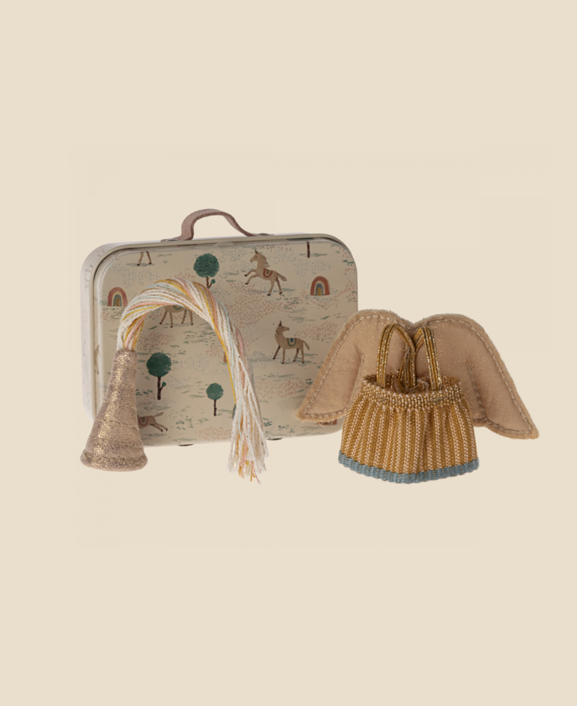 A Maileg Pig in a Box with Fairy Outfit, featuring a horse and rainbow patterned suitcase, angel wings, a gold beaded bag, and a tassel party hat are displayed on a plain beige background.
