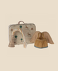 The Maileg Little Sister on Rocking Horse Set is pictured with a small suitcase featuring a whimsical animal and rainbow print, fairy clothes, and a gold tassel party hat—all arranged on a pale background for a magical scene.