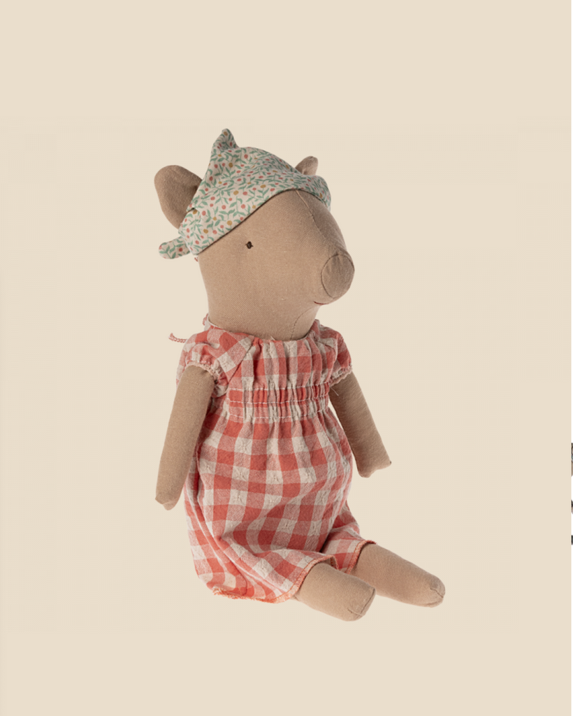 A Maileg Pig - Girl sits upright against a plain background. The girl pig is dressed in a red and white checkered dress and wears a floral-patterned headscarf. Its expression is neutral, and it appears to be handmade with stitched details.