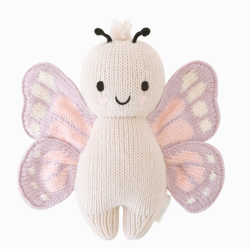 A Cuddle + Kind Baby Butterfly made from Peruvian cotton yarn, featuring a white body, light purple wings with pink and white accents, black stitched eyes, a smiling mouth, and two short black antennae atop its head. This collectible piece sits against a plain white background.