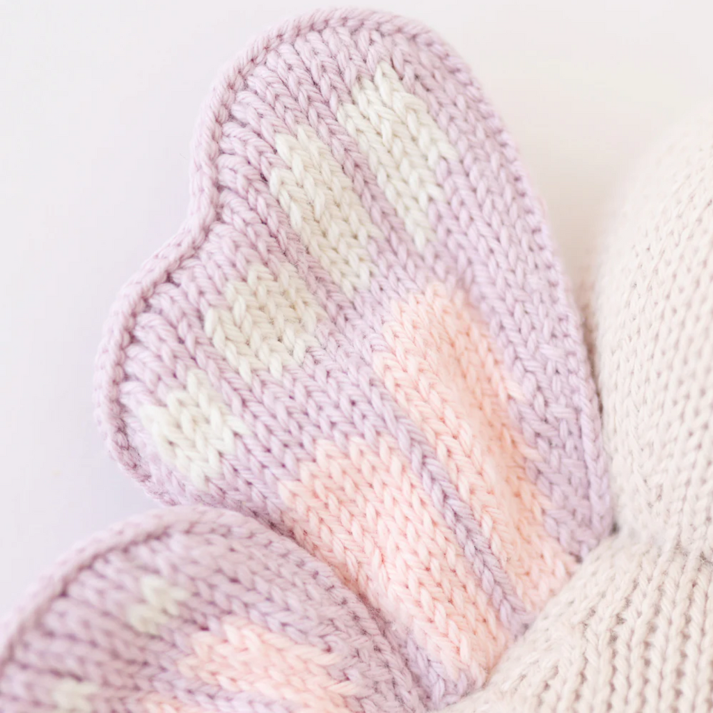Close-up of a knitted butterfly wing in pastel colors. The wing, crafted from soft Peruvian cotton yarn, features a heart-shaped lilac border with a pattern of white and pink stitches on a light purple background. This delicate piece is perfect for any collection of Cuddle + Kind Baby Butterfly or other keepsakes.