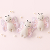 Three cute Cuddle + Kind Baby Butterfly plush toys made from Peruvian cotton yarn on a light pink background. Each plush has a cream-colored body with black antennas and wings in pastel shades of purple and pink. Small white flowers are scattered around the heirloom-quality toys.