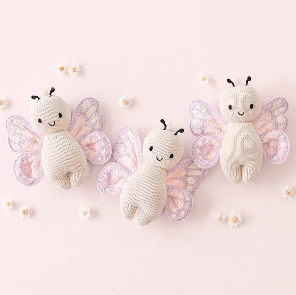 Three cute Cuddle + Kind Baby Butterfly plush toys made from Peruvian cotton yarn on a light pink background. Each plush has a cream-colored body with black antennas and wings in pastel shades of purple and pink. Small white flowers are scattered around the heirloom-quality toys.