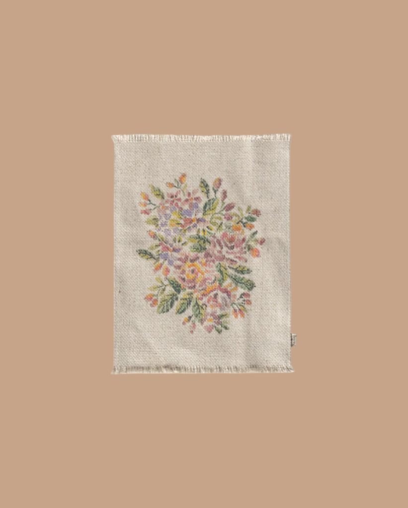 A cream, woven textile with frayed edges showcases a beautiful floral embroidery in pink, purple, yellow, and green. This design echoes the charm of a Maileg Flower Rug on its light brown background.
