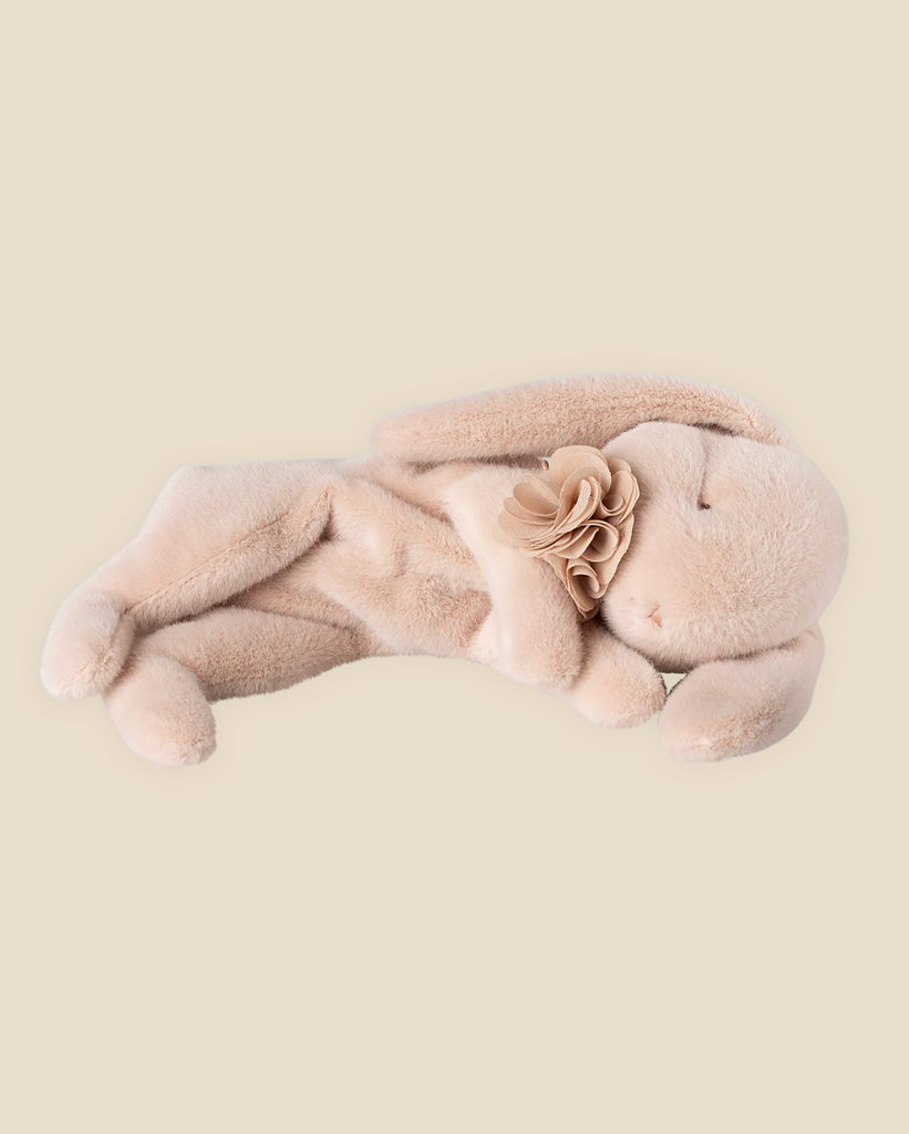 A Maileg Plush Sleeping Bunny - Powder, crafted from soft cotton fabric, lies on its side with long ears and eyes gently closed. It features a small flower decoration near its neck, set against a plain beige background. This charming piece ships in one week.