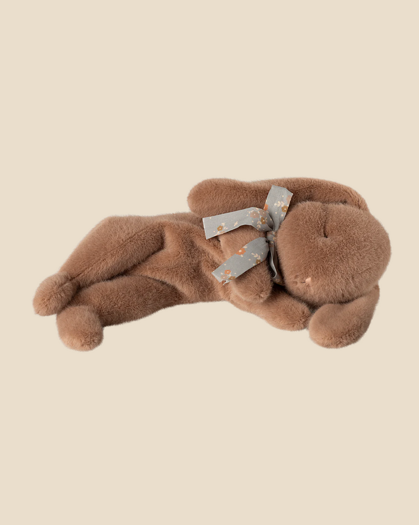 The Maileg Plush Sleeping Bunny in soft brown, with floppy ears and stitched eyes, lies on its side like a sleeping bunny against a plain background. It's crafted from Maileg cotton fabric and adorned with a gray ribbon featuring a small floral pattern around its neck, creating an irresistibly cuddly look.