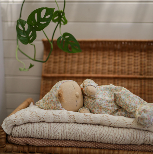 An Olli Ella Dozy Dinkums - Custard - Garden Floral doll in floral pajamas and cap rests on folded knitted blankets. A wicker chest and potted plant with vibrant leaves complete the background, making it an ideal Easter companion.