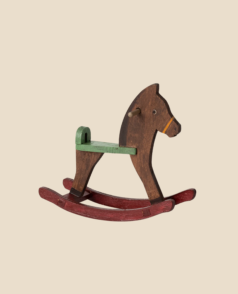 The Maileg Mouse Rocking Horse, featuring a vintage brown body, green seat, and red rockers, elegantly rests on a beige backdrop. Its classic design evokes nostalgic memories of timeless wooden toys. (Ships in one week).