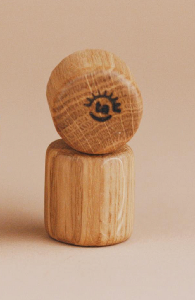 A wooden figurine with a round head and cylindrical body stands upright, about the size of a Maileg Moms Afternoon Tea Set. Its simply drawn winking face smiles against a soft beige background.