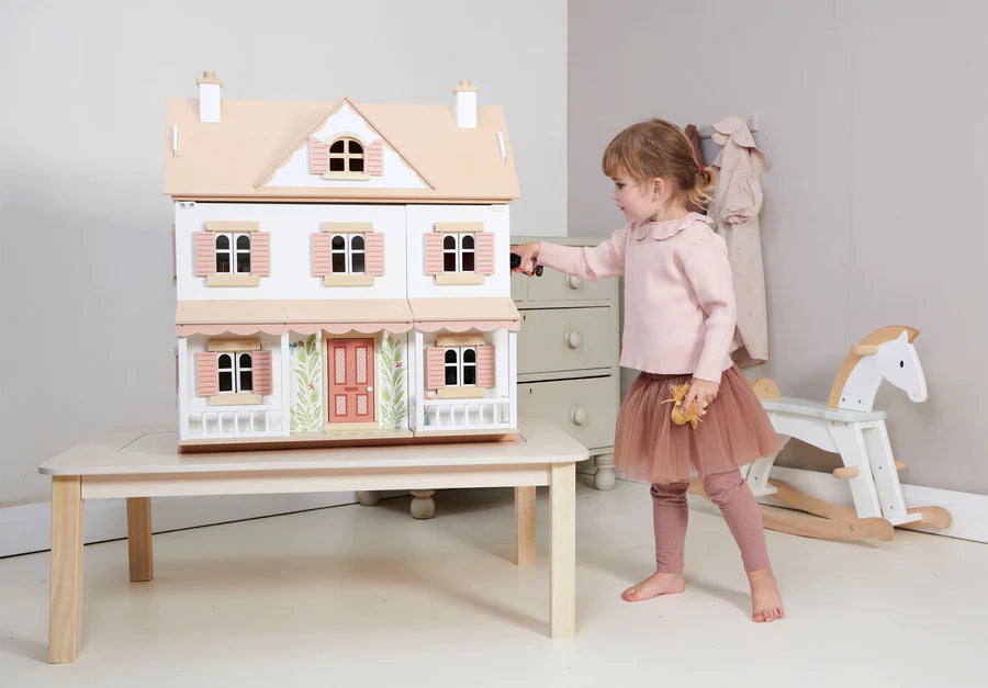 A young girl wearing a pink top and a brown tutu stands next to a dollhouse on a table. The detailed Humming Bird Dollhouse, with its white exterior, pink shutters, and red door, appears as if it belongs to an exquisite furniture collection. A white rocking horse is in the background, adding to the room's light and airy feel.