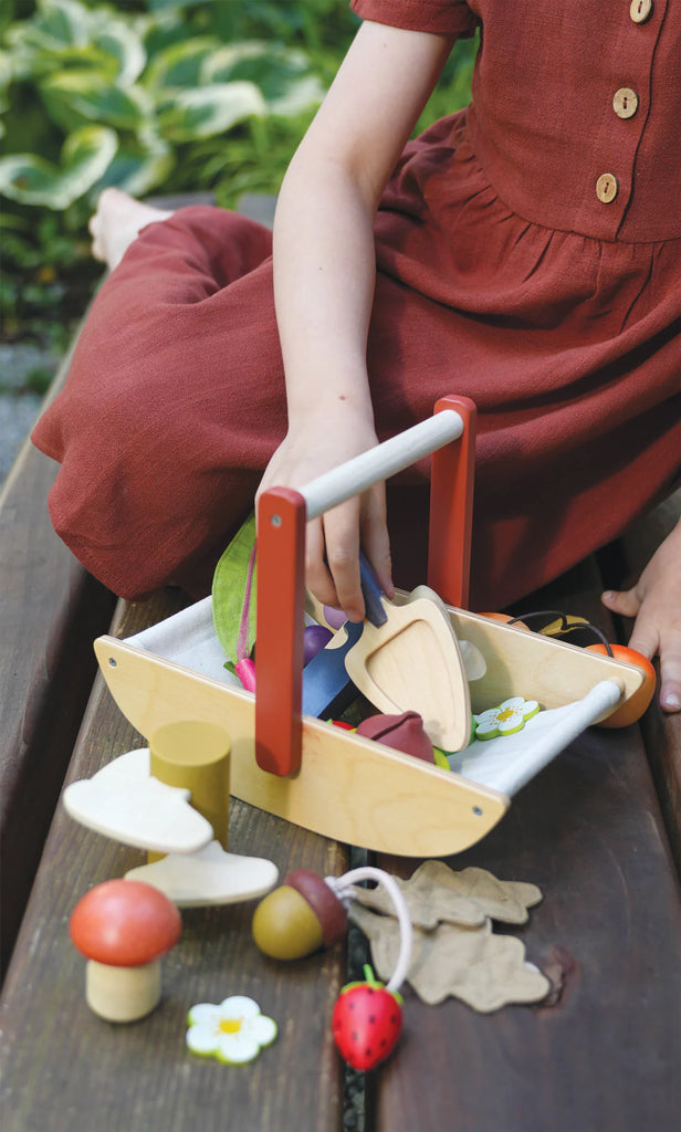 A child in a brown dress plays on a wooden surface with the Wild Wood Foraging Trug, filled with colorful toy vegetables and flowers. Rummaging through the trug amidst greenery, it's as if they've collected foraged plants for an imaginative feast.