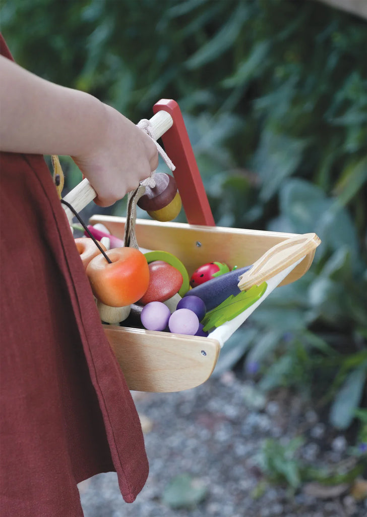 Wearing a brown apron, someone holds the Wild Wood Foraging Trug filled with toy fruits and vegetables, including an apple, eggplant, and carrots, amidst a garden backdrop of lush green plants.