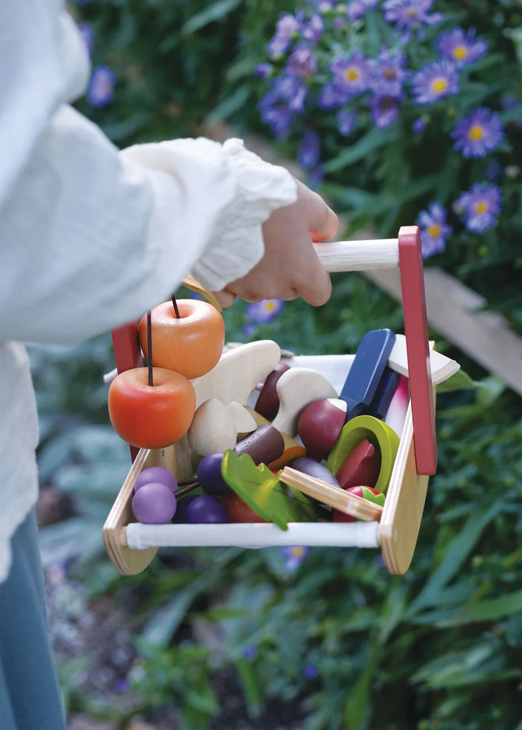 Someone holds a Wild Wood Foraging Trug, filled with an assortment of colorful toy fruits and vegetables, reminiscent of foraged plants. The basket's red handles stand out against a lush backdrop of purple flowers and green foliage, enhancing the whimsical scene.