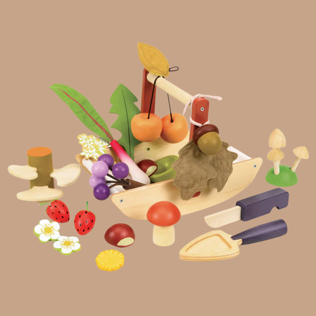 The Wild Wood Foraging Trug features a wooden toy set with fruits, vegetables, and gardening tools on a brown background. It includes cherries, grapes, mushrooms, foraged plants, a handled basket, and a small shovel to inspire kids in imaginative play.