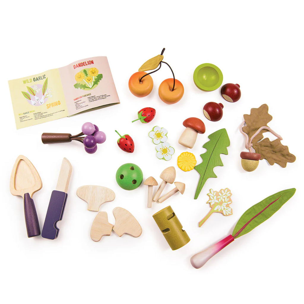 The Wild Wood Foraging Trug features a collection of wooden toy foods, including a sliceable peach, mushrooms, cherries, strawberries, and leafy greens. It also includes small books on foraged plants like wild garlic and dandelion, plus a toy knife and cutting board.