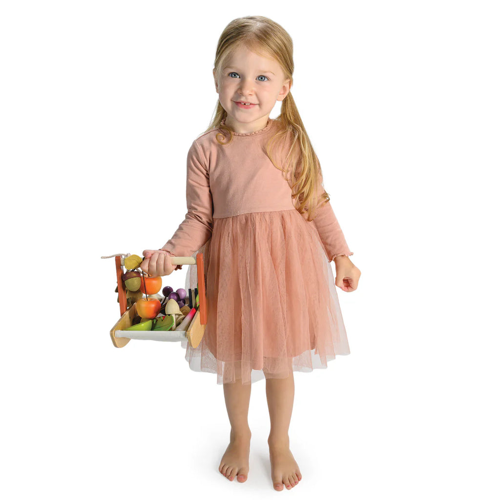 Barefoot, a young girl in a peach dress beams as she holds the Wild Wood Foraging Trug, filled with colorful toy fruits and vegetables. A whimsical recipe leaflet inside hints at fun culinary adventures against a pristine white backdrop.