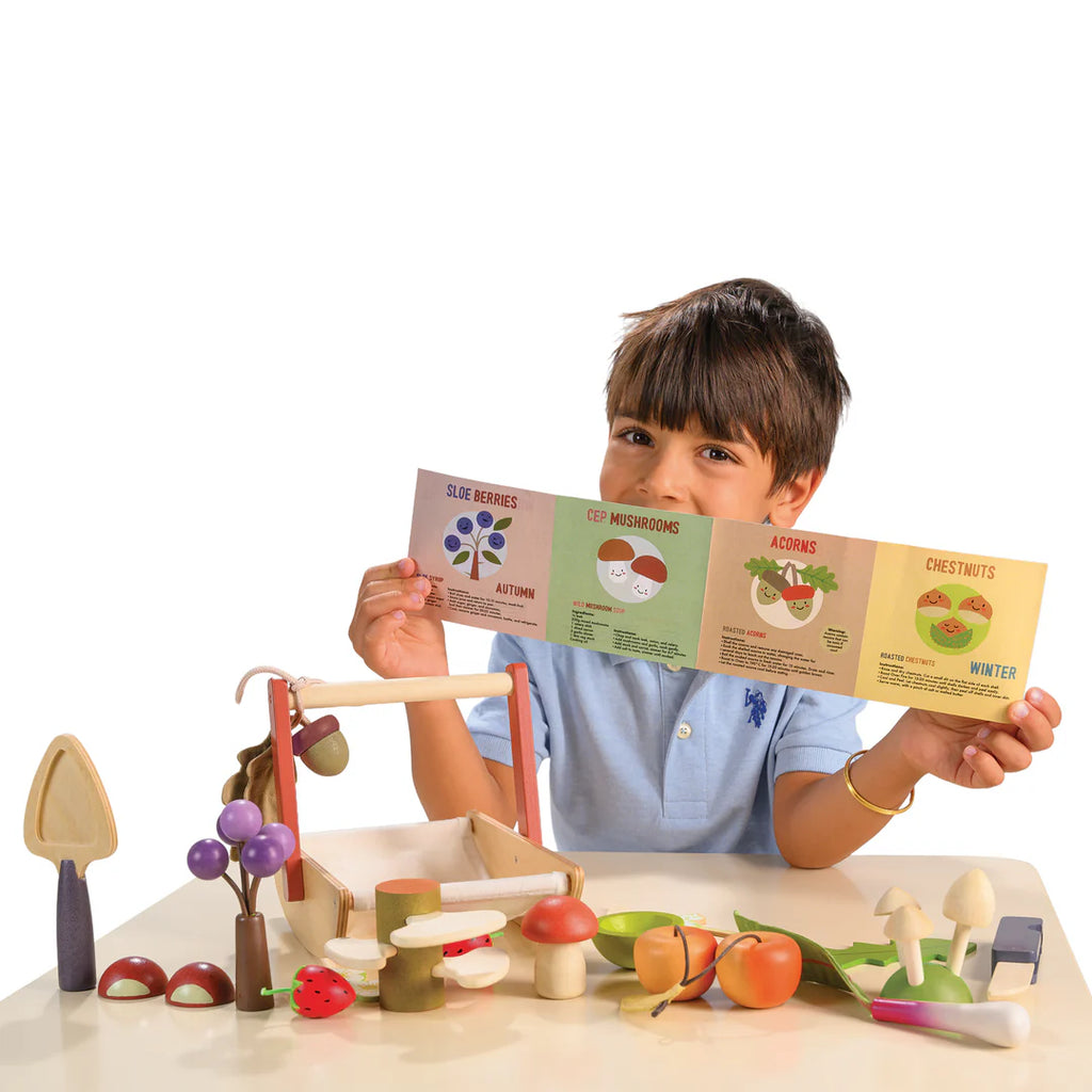 A child sits with an educational booklet about seasonal foods, surrounded by a Wild Wood Foraging Trug filled with colorful wooden toy mushrooms, berries, and vegetables. Engaged and smiling, the child imagines foraged plants adding magic to their interactive learning.