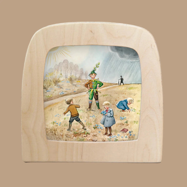 Children play in a flowered field under a split sky, half sunny and half stormy. A man in green casts Magic Silhouettes at the center, with another translucent figure nearby. The scene is framed in a rounded wooden square for the Silhouette Only - Around The Year (April) for Toverlux Lamp.