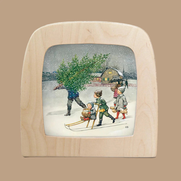 A vintage illustration shows a snowy scene with a person carrying a Christmas tree and three children on a sled. A warmly lit house glows like the Silhouette Only - Around The Year (December) for Toverlux Lamp, beautifully framed by a wooden border.