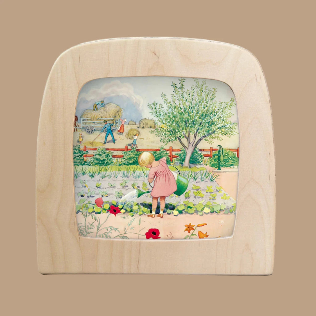 The "Silhouette Only - Around The Year (July) for Toverlux Lamp" features a wooden picture frame showcasing a child in a pink dress watering plants, with translucent material depicting farmers by hay bales beneath a cloudy sky, framed by a tree and red fence.