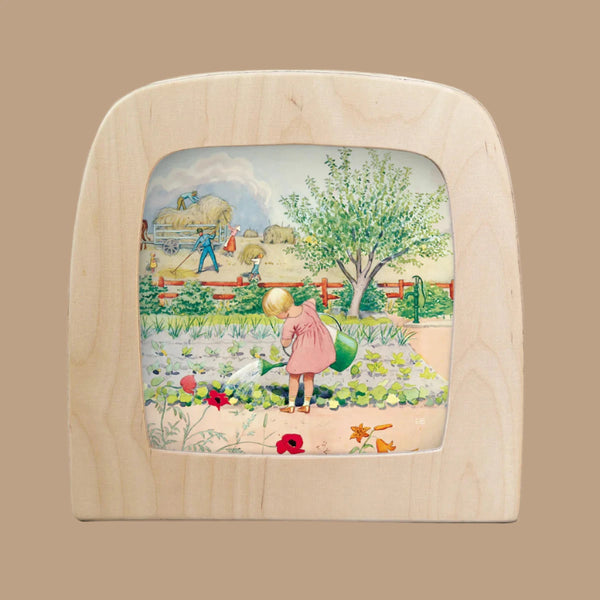 The "Silhouette Only - Around The Year (July) for Toverlux Lamp" features a wooden picture frame showcasing a child in a pink dress watering plants, with translucent material depicting farmers by hay bales beneath a cloudy sky, framed by a tree and red fence.
