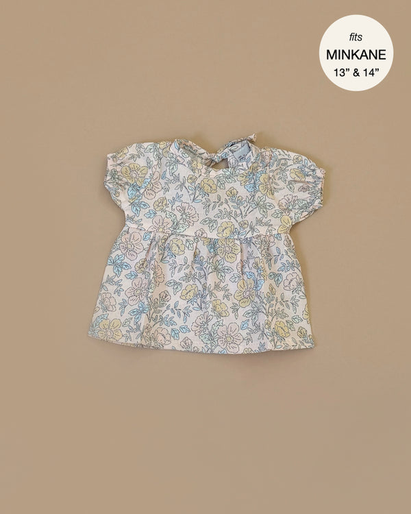 The Faustine dress with balloon sleeves Celia, a floral baby doll dress for Minikane babies, is laid flat against a beige background. With light fabric and shades of blue, yellow, and pink flowers, it features a label reading "fits Minikane 13'' & 14''.