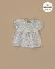 The Faustine dress with balloon sleeves Celia, a floral baby doll dress for Minikane babies, is laid flat against a beige background. With light fabric and shades of blue, yellow, and pink flowers, it features a label reading "fits Minikane 13'' & 14''.