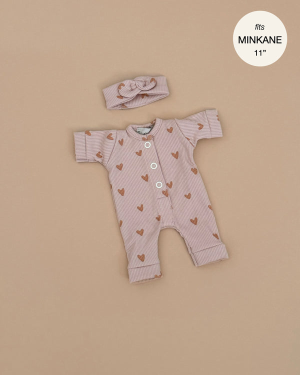 The Minikane Doll Clothing Lili Jumpsuit, set against a beige background, is perfect for Minikane dolls. It includes a French-made long-sleeve onesie with ribbed pink heart patterns and white buttons, paired with a matching headband and bow. A circular label reads "fits MINIKANE 11.”.