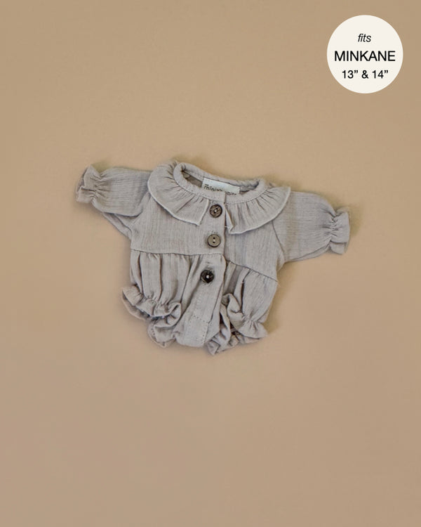 The Leonore Romper in Pebble Cotton Gauze by Minikane is a small, light brown, long-sleeve romper with buttons and a frilled collar for hand washable dolls. It’s displayed on a beige background with a circular label stating "fits MINKANE 13” & 14”" in the top right corner.