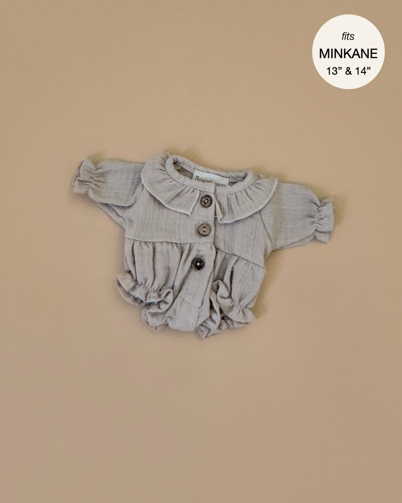The Leonore Romper in Pebble Cotton Gauze by Minikane is a small, light brown, long-sleeve romper with buttons and a frilled collar for hand washable dolls. It’s displayed on a beige background with a circular label stating "fits MINKANE 13” & 14”" in the top right corner.