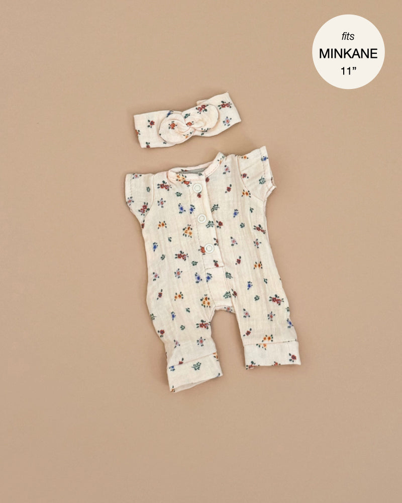 The Lili Jumpsuit in Elise Cotton Gauze, a cream floral-patterned doll outfit complete with buttons and matching headband, is showcased on a beige backdrop. Designed for Minikane dolls, it features the label "fits MINKANE 11," and is made in France. Hand washable for easy care.