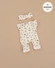 The Lili Jumpsuit in Elise Cotton Gauze, a cream floral-patterned doll outfit complete with buttons and matching headband, is showcased on a beige backdrop. Designed for Minikane dolls, it features the label "fits MINKANE 11," and is made in France. Hand washable for easy care.