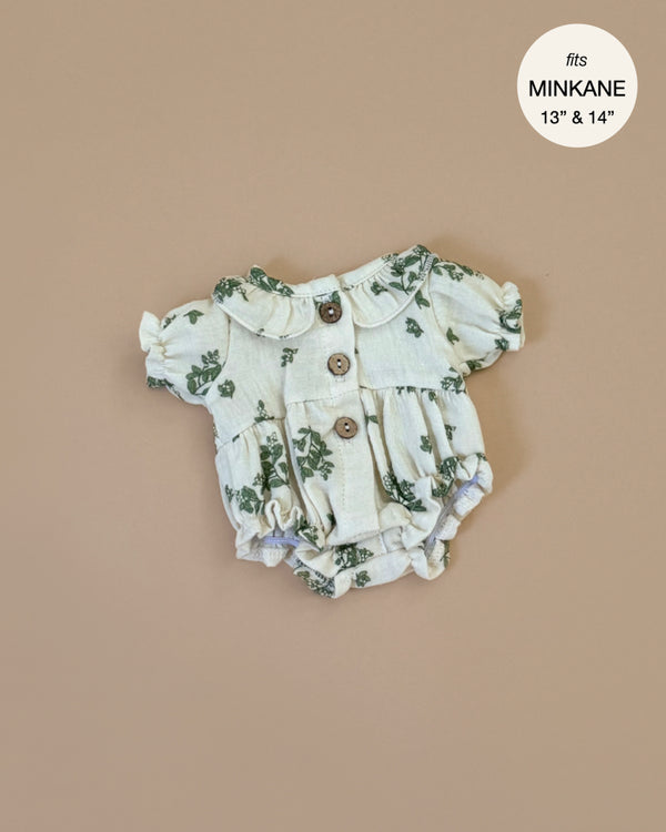 The Leonore Romper in Green Foliage for Minikane dolls is showcased on a beige background. This cream romper has a green floral design, short puffed sleeves, and wood-look buttons. A label says "fits MINKANE 13” & 14” dolls," emphasizing its hand washable fabric.