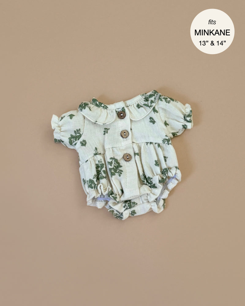 The Leonore Romper in Green Foliage for Minikane dolls is showcased on a beige background. This cream romper has a green floral design, short puffed sleeves, and wood-look buttons. A label says "fits MINKANE 13” & 14” dolls," emphasizing its hand washable fabric.