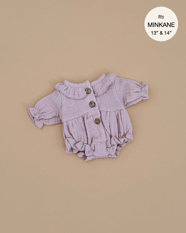 The Leonore Romper for Minikane Gordis dolls is a lavender, long-sleeve piece with a ruffled collar and three front buttons. Made in France from petal cotton gauze, it fits 13'' & 14'' dolls and is hand washable. It’s displayed on a beige background.