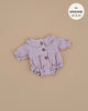 The Leonore Romper for Minikane Gordis dolls is a lavender, long-sleeve piece with a ruffled collar and three front buttons. Made in France from petal cotton gauze, it fits 13'' & 14'' dolls and is hand washable. It’s displayed on a beige background.