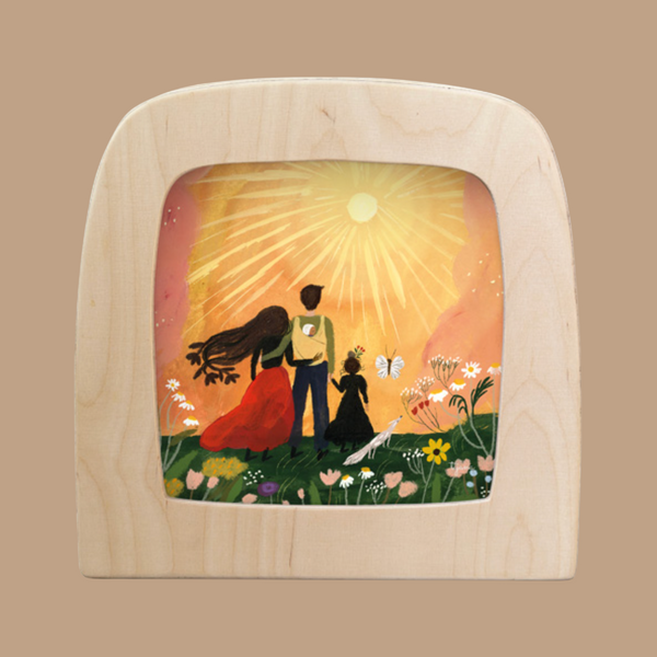 The Silhouette Only - Summer Solstice for Toverlux Lamp features a wooden Magic Window Frame with an illustration of a family, including an adult, child, and long-haired figure, standing in a flower-filled field under the bright sun.