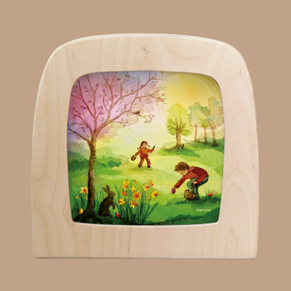 The "Silhouette Only - Easter Discoveries for Toverlux Lamp" features a wooden frame with an illustration of two children in a vibrant spring landscape, one picking flowers near daffodils and the other skipping under a blooming tree, capturing the magic of silhouettes.