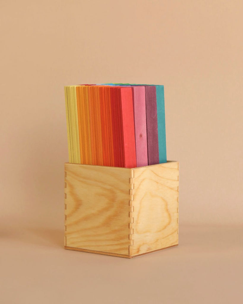 A wooden box contains neatly organized vertical dividers, each in a different color ranging from yellow, orange, red, pink, to blue. The background is a soft beige, complementing the natural wood texture of the box—perfect for fine motor skills development akin to Grimm's Leonardo da Vinci Building Sticks.