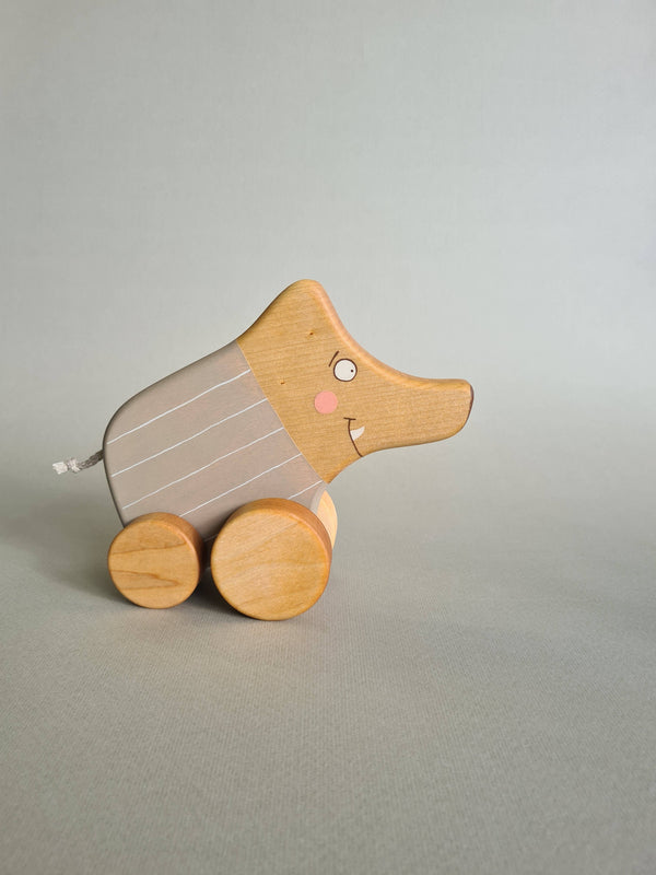 The Pull and Push Toy Little Wild Boar is a handcrafted wooden toy featuring a curving form with light gray paint, white stripes, and rests on a light green background, highlighting the charm of sustainable materials.