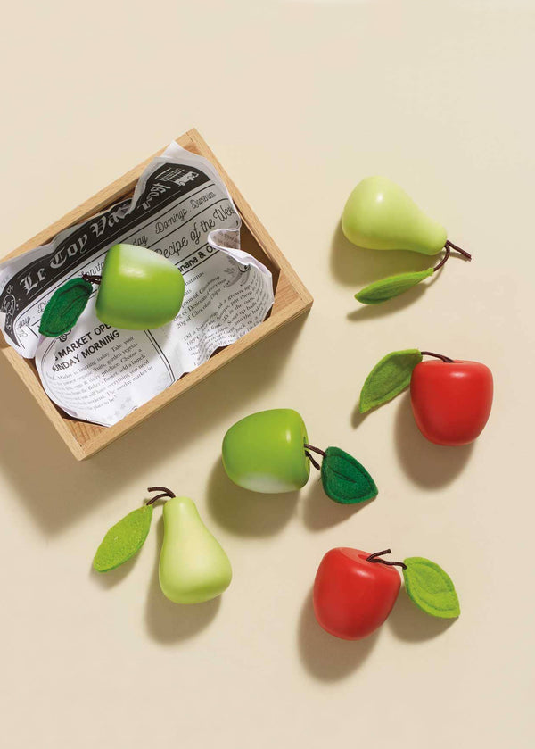 The Orchard Fruits Wooden Market Crate includes a newspaper-lined wooden box with a green pear. Nearby are four fruit sculptures: a green pear, two wooden apples and pears, and a red apple, all with green leaves on a pale yellow surface resembling an early-learning toy set.