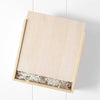 A wooden gift box with a sliding lid, partially open, reveals shredded white packing material holding the Newborn Gift Set. The pristine white background enhances the simple elegance of the box, hinting at treasures for both baby and parents.