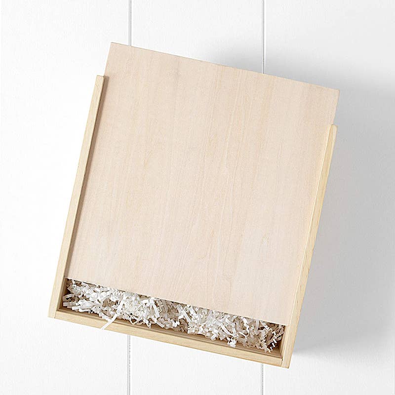 A wooden gift box with a sliding lid, partially open, reveals shredded white packing material holding the Newborn Gift Set. The pristine white background enhances the simple elegance of the box, hinting at treasures for both baby and parents.
