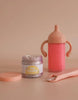 A small jar from the "My tiny birthday set - Tiny Harlow," resembling a birthday cake jar, sits with a pink sippy cup and spoon on a beige surface. The open lid invites little ones into the world of realistic play.
