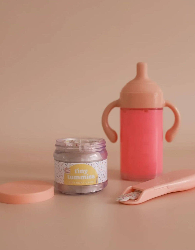 A small jar from the "My tiny birthday set - Tiny Harlow," resembling a birthday cake jar, sits with a pink sippy cup and spoon on a beige surface. The open lid invites little ones into the world of realistic play.